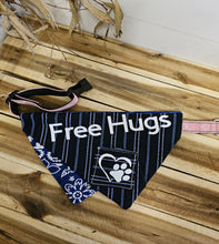 Load image into Gallery viewer, A navy blue and white striped dog bandana with 'Free Hugs' text and a paw print graphic, shown on a rustic wooden background with a pink and black leash attached.