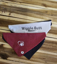 Load image into Gallery viewer, Two triangular dog bandanas laid flat on a wooden surface: one white with black text reading 'Wiggle Butt' and one red-and-white gingham with a white paw print logo and small circular 'S' mark. No person or animal is present.