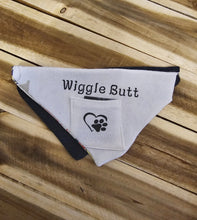 Load image into Gallery viewer, A white triangular pet bandana with black trim, featuring the text 'Wiggle Butt' and a black paw print logo, laid flat on a wooden plank surface.