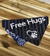 Load image into Gallery viewer, A navy blue pet bandana with white stripes, featuring the text 'Free Hugs' in white, a paw print in a cage graphic, and a matching blue and white floral patterned corner, laid flat on a wooden plank background.
