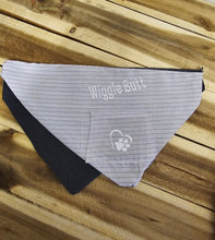 Load image into Gallery viewer, A triangular pet bandana laid flat on a wooden surface, featuring a light gray and white striped front panel with the words 'Wiggle Butt' and a small paw-print heart patch, plus a dark navy backing.