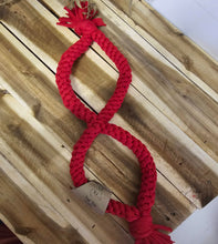 Load image into Gallery viewer, A red braided rope pet toy shaped like an infinity symbol, laid diagonally on a light brown wooden plank background. A small paper tag is attached near one end.