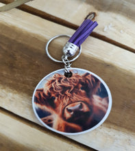 Load image into Gallery viewer, Keychain with a circular tag featuring a lCow design on a wooden surface