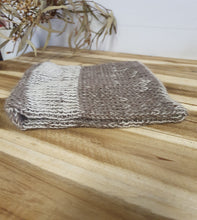 Load image into Gallery viewer, Knitted gray and white dishcloth hanging on a wooden surface with a blurred plant in the background.