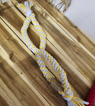 Load image into Gallery viewer, A braided dog toy made of yellow and white rope with tassel ends, laid diagonally on a wooden plank floor.