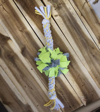 Load image into Gallery viewer, A braided dog toy made from white, yellow, and gray fabric, featuring a central cluster of green leaf-shaped pieces, displayed on a wooden plank surface.