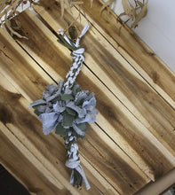 Load image into Gallery viewer, A natural dog chew toy made of braided white and tan cowhide with a central cluster of dusty sage green leaves, laid diagonally on a rustic wooden plank surface.

