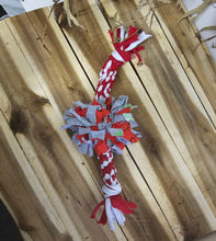 Load image into Gallery viewer, A red, white, and gray fabric dog chew toy with knotted ends, displayed diagonally on a light brown wooden plank surface.
